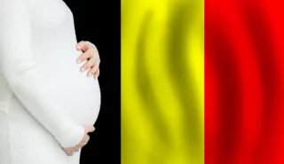 Pregnant woman in a white knit sweater cradling her belly, with the Belgian flag's black, yellow, and red vertical stripes in the background.