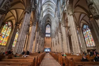 Interior of a Gothic cathedral with tall columns, pointed arches, and colorful stained-glass windows, and visitors in pews.