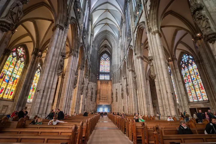 Interior of a Gothic cathedral with tall columns, pointed arches, and colorful stained-glass windows, and visitors in pews.
